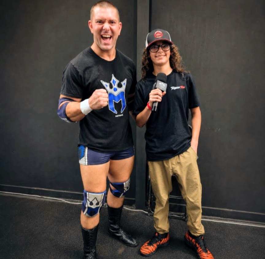 Wrestler Mathias flexing beside Trystan of Turnbuckle Tribe, who holds a microphone, at CCP Podcasting in Casselberry, Florida