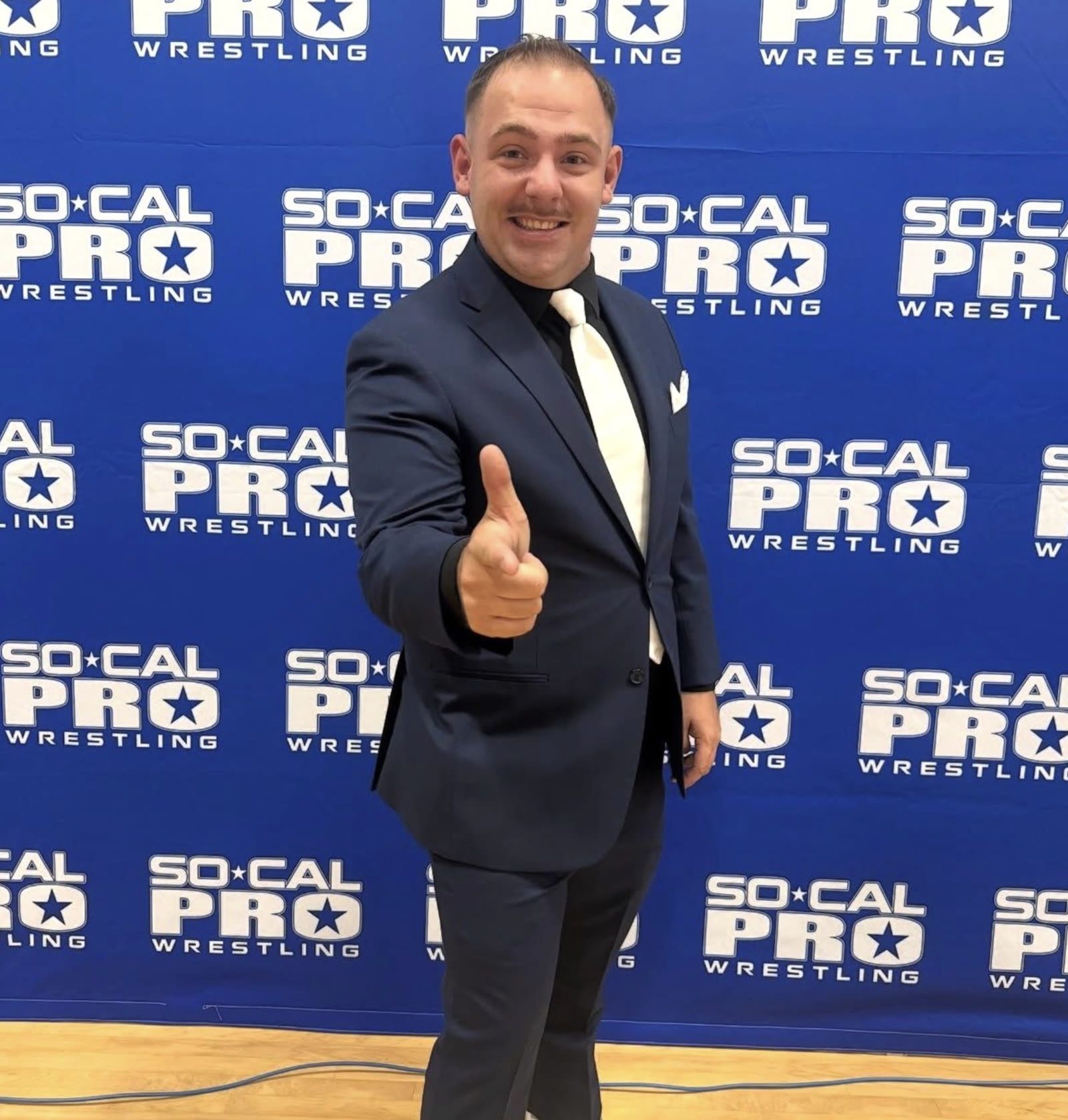 Kevin Cormac giving a thumbs up in a navy suit in front of a SoCal Pro Wrestling backdrop