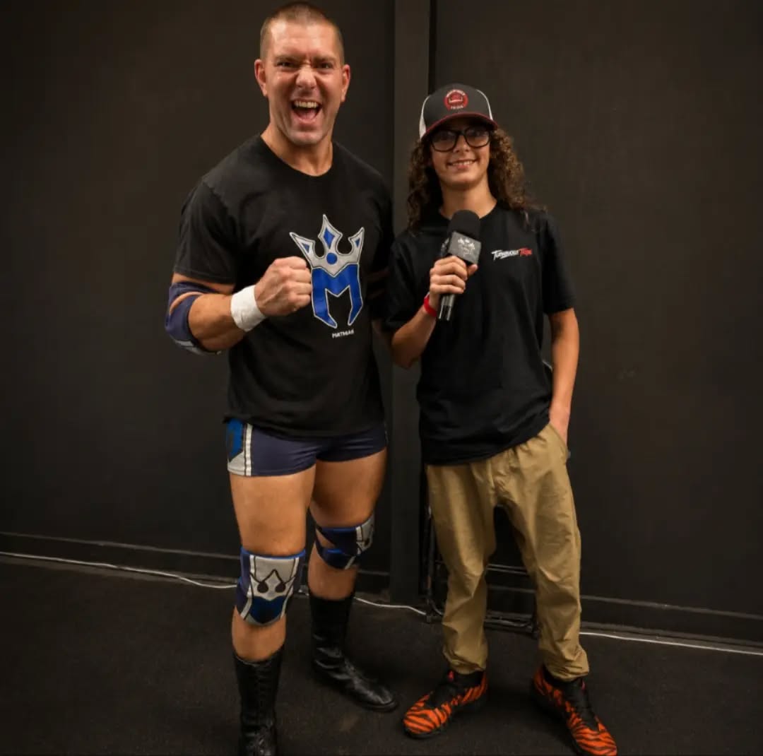 Trystan holds a microphone beside a professional wrestler