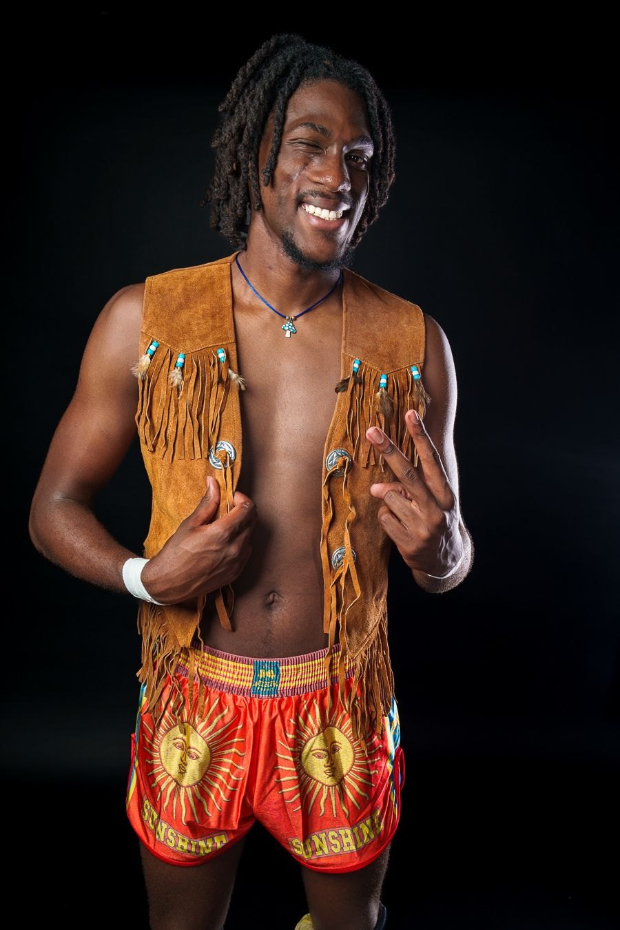 Gage Holloway, the Midwest Bluebird, winking and flashing a peace sign in a fringed suede vest and orange sunshine trunks